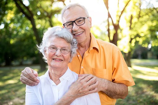 An elderly couple with glasses stand outside with the mans arms around the woman's shoulders as they smile
