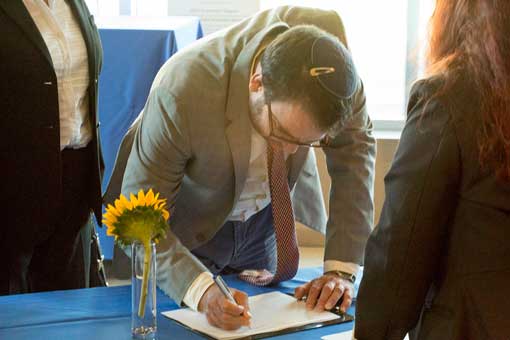 A man wearing a yamaka leans over signing a paper